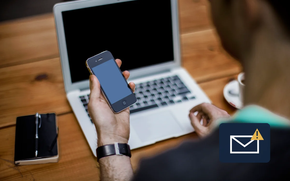 Illustrative photograph of a person using a phone beside a laptop, with an email-warning symbol, for account security advice.